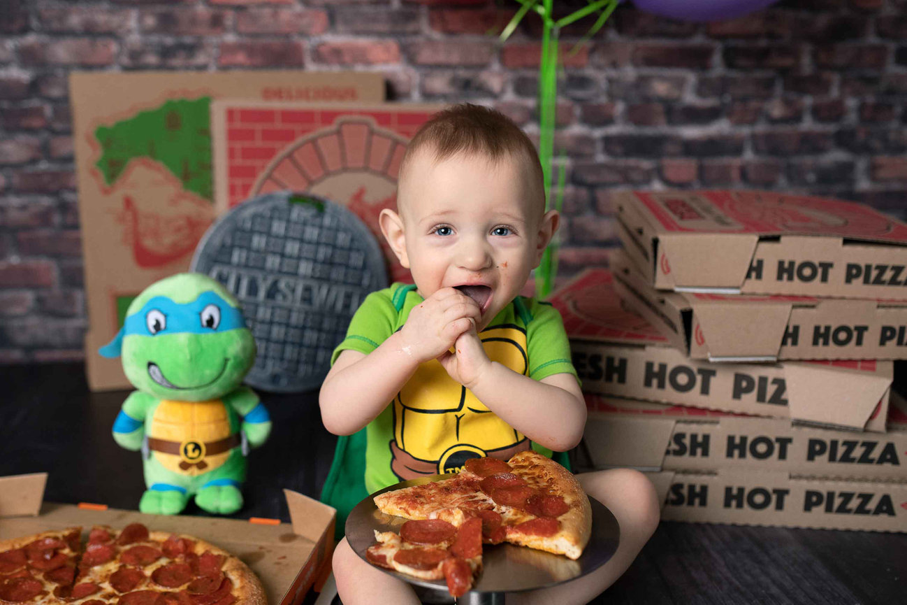 A baby dressed in a turtle costume smiles amid pizza boxes in Lake in the Hills, IL. Holding a slice of pepperoni pizza, the child sits beside a plush turtle toy. A brick wall with a cityscape motif provides an urban backdrop to this charming scene.