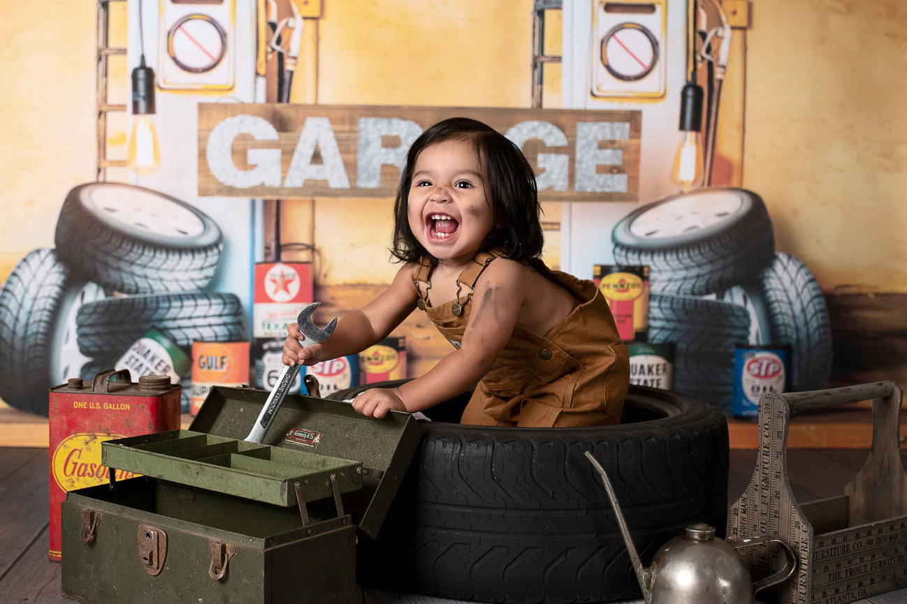A joyful toddler celebrating a baby birthday, wearing brown overalls, sits in a tire surrounded by vintage garage items. The child is holding a wrench. In the background, stacked tires and a GARAGE sign set the scene for this milestone moment.