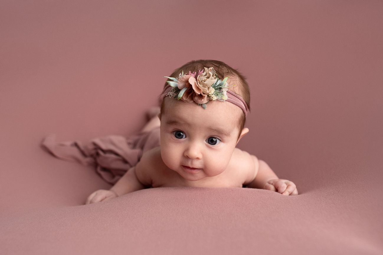 A baby lies on a soft, light pink surface, wearing a pink headband with floral decorations. The baby gazes forward with wide eyes and a gentle expression, capturing the essence of portrait photography in its purest form.