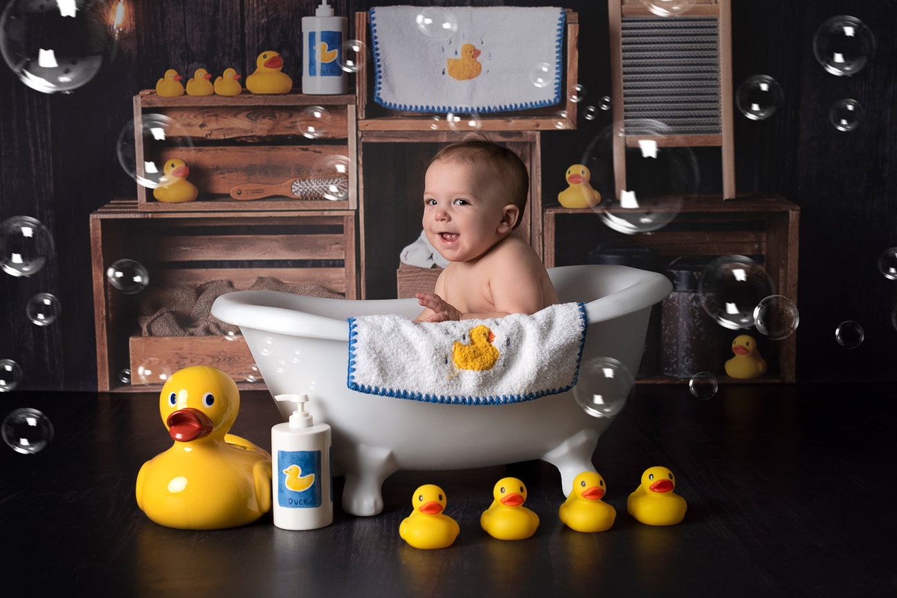 A 9-month-old baby sits smiling in a white clawfoot tub, surrounded by yellow rubber ducks and bubbles. The background features shelves with duck-themed decorations, capturing the essence of joyful childhood.