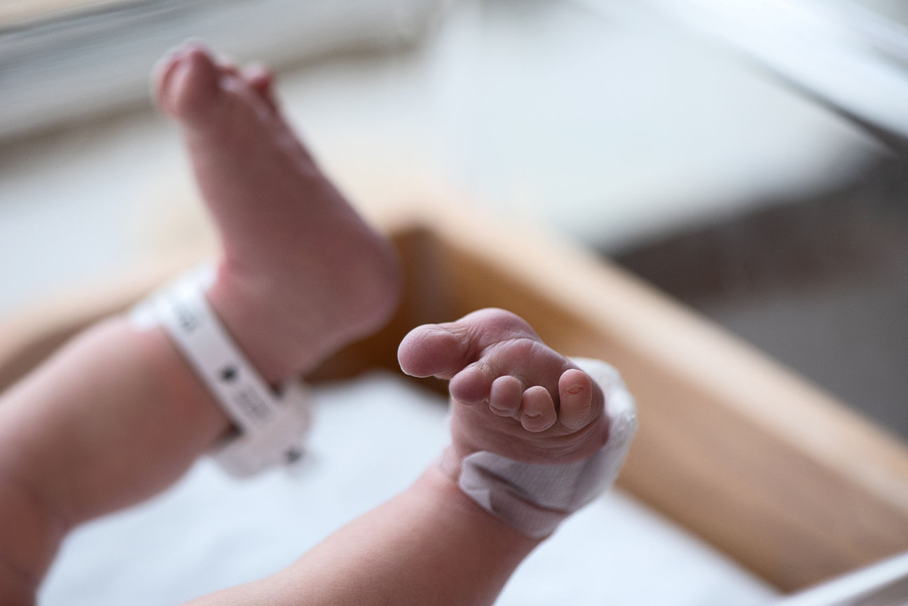 Close-up of a newborns feet at an Illinois hospital, one leg adorned with a hospital ID band and the other with a medical bandage. The baby rests on a soft surface, with part of a Barrington crib visible in the blurred background.