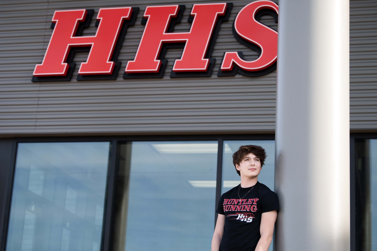 A senior from Huntley, Illinois, proudly sports a Huntley Running T-shirt while leaning against a pillar outside a building with large red letters HHS above the entrance. The structure features sleek glass windows set in a grid-patterned facade.