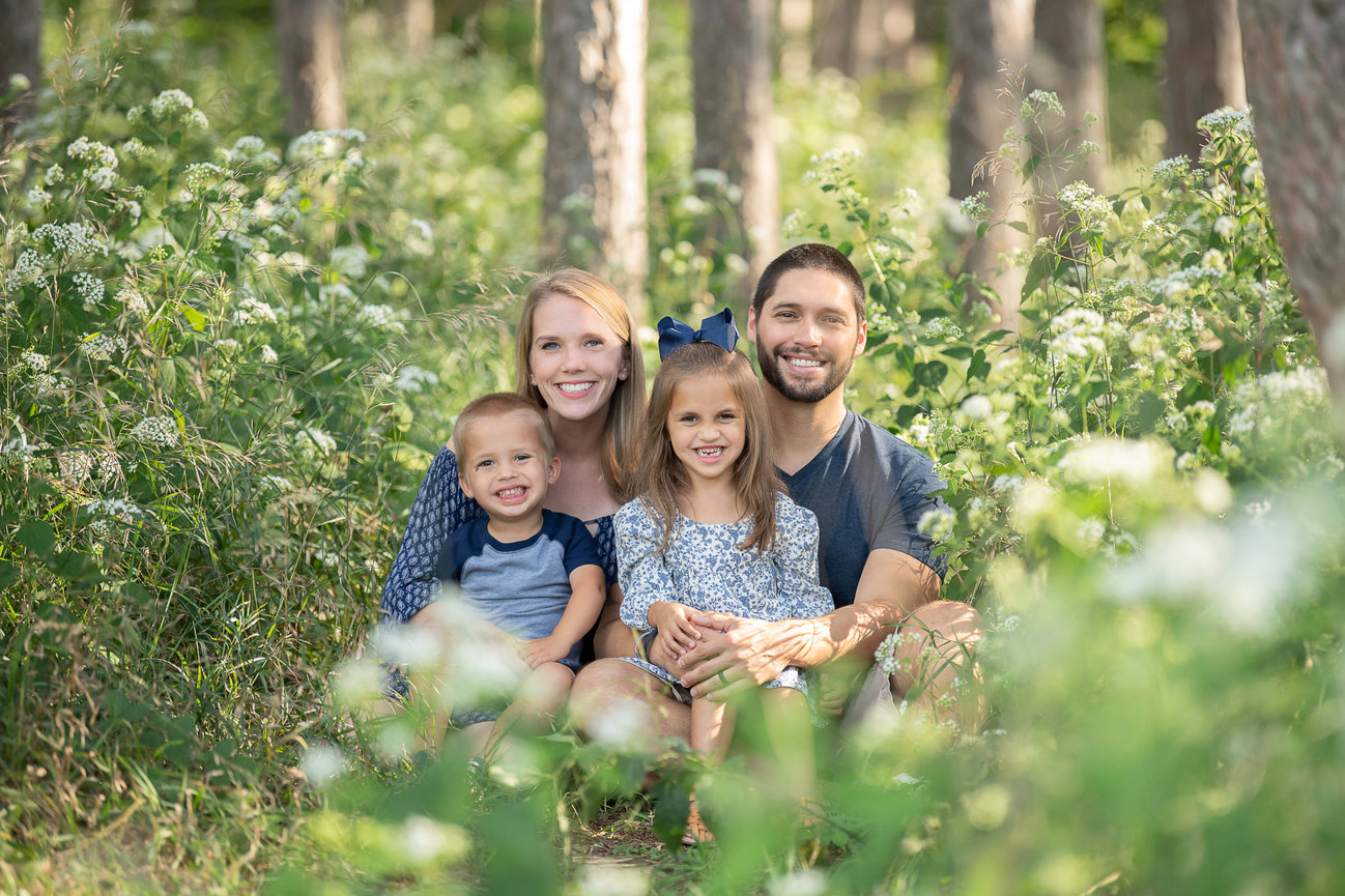 A family of four, consisting of two adults and two children, pose happily among tall trees and lush greenery. Captured by Crystal Lake Photography, the parents sit with their daughter and son, all smiling in a sunlit setting surrounded by wildflowers