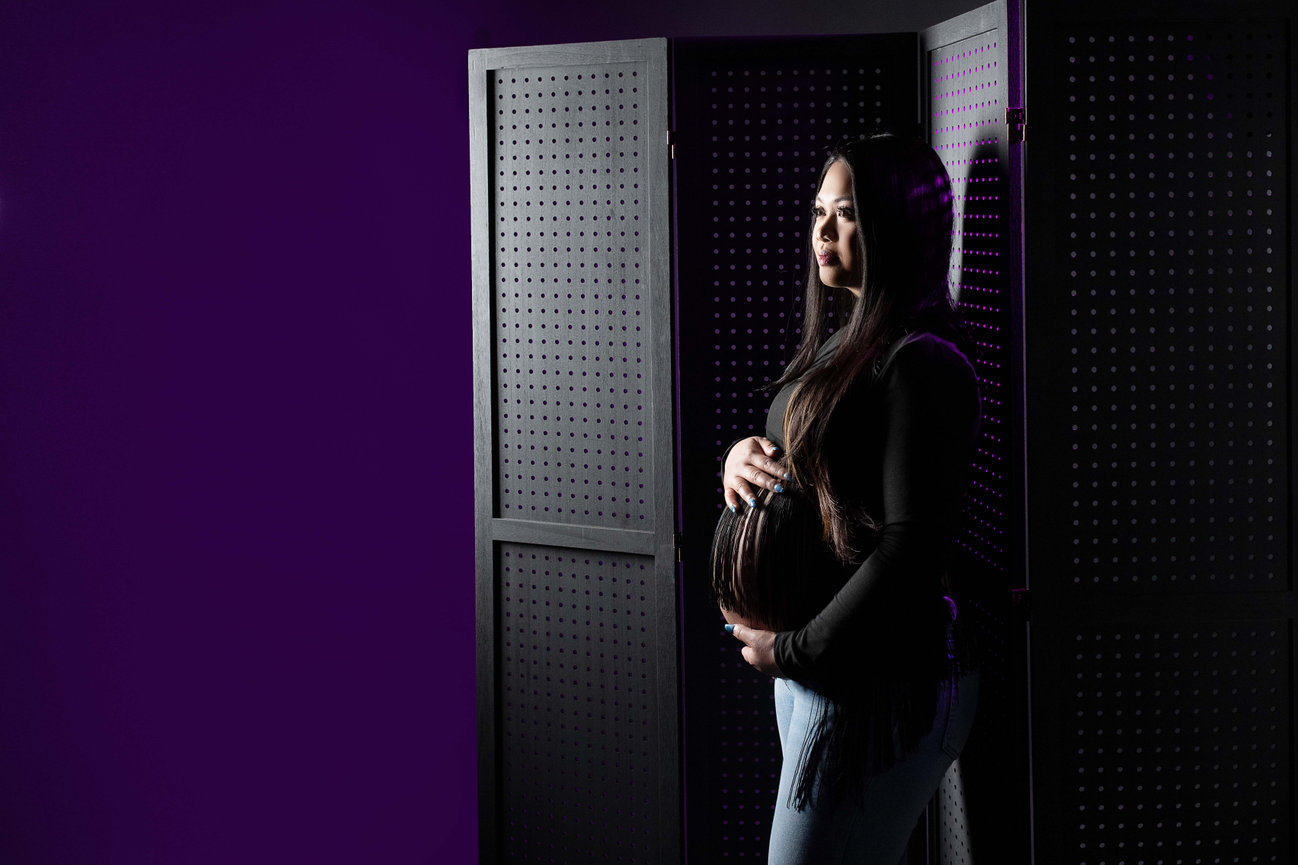 A pregnant woman stands in front of a perforated privacy screen, set against a purple backdrop reminiscent of twilight over Crystal Lake in Illinois. Her long dark hair cascades over a black top as she gently cradles her belly with both hands.