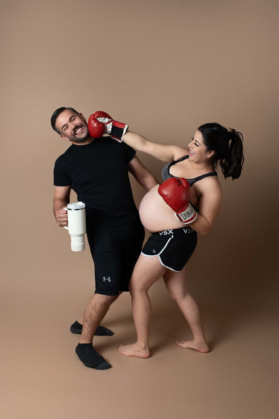 A pregnant woman playfully punches a man with boxing gloves. Shes wearing shorts and a sports bra, while the man holds a large mug and wears a black t-shirt and shorts. They both appear to be having fun against a neutral background.