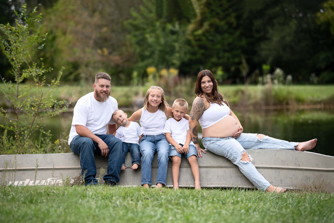 A family of five sits on a metal boat by a pond. A pregnant woman with tattoos rests her legs on the edge. Two children, a girl and a boy, sit next to her, and a man holds a toddler on his lap. All wear casual jeans and white tops.