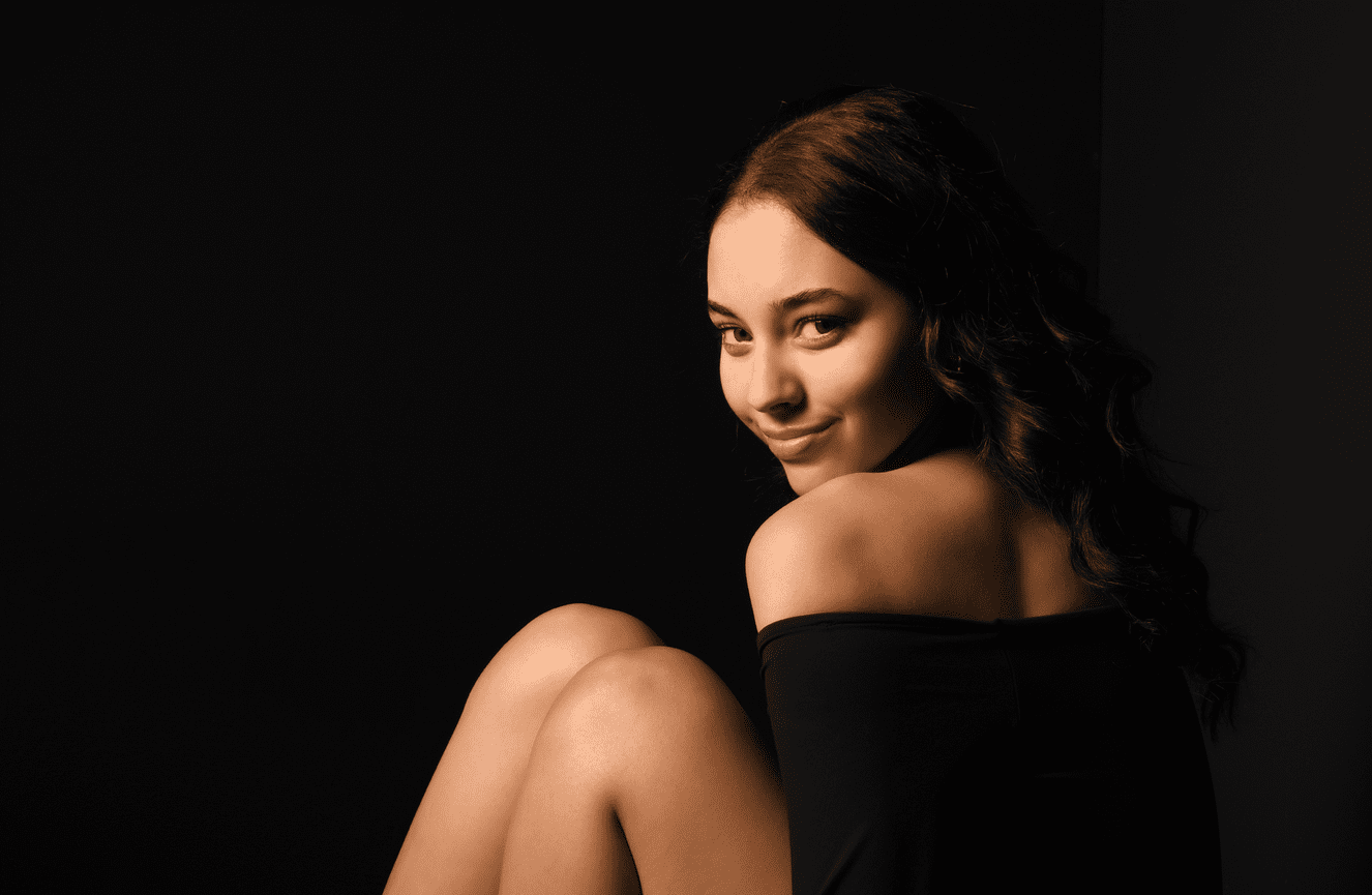 A brunette girl wearing an off the shoulder dress looks over her shoulders with her knees curled up to her chest.