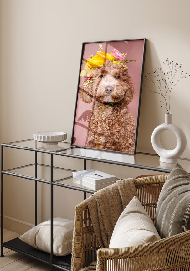 A framed portrait of an apricot cavoodle wearing a flower crown against a dusty pink background, displayed on a glass console table beside a vase and books
