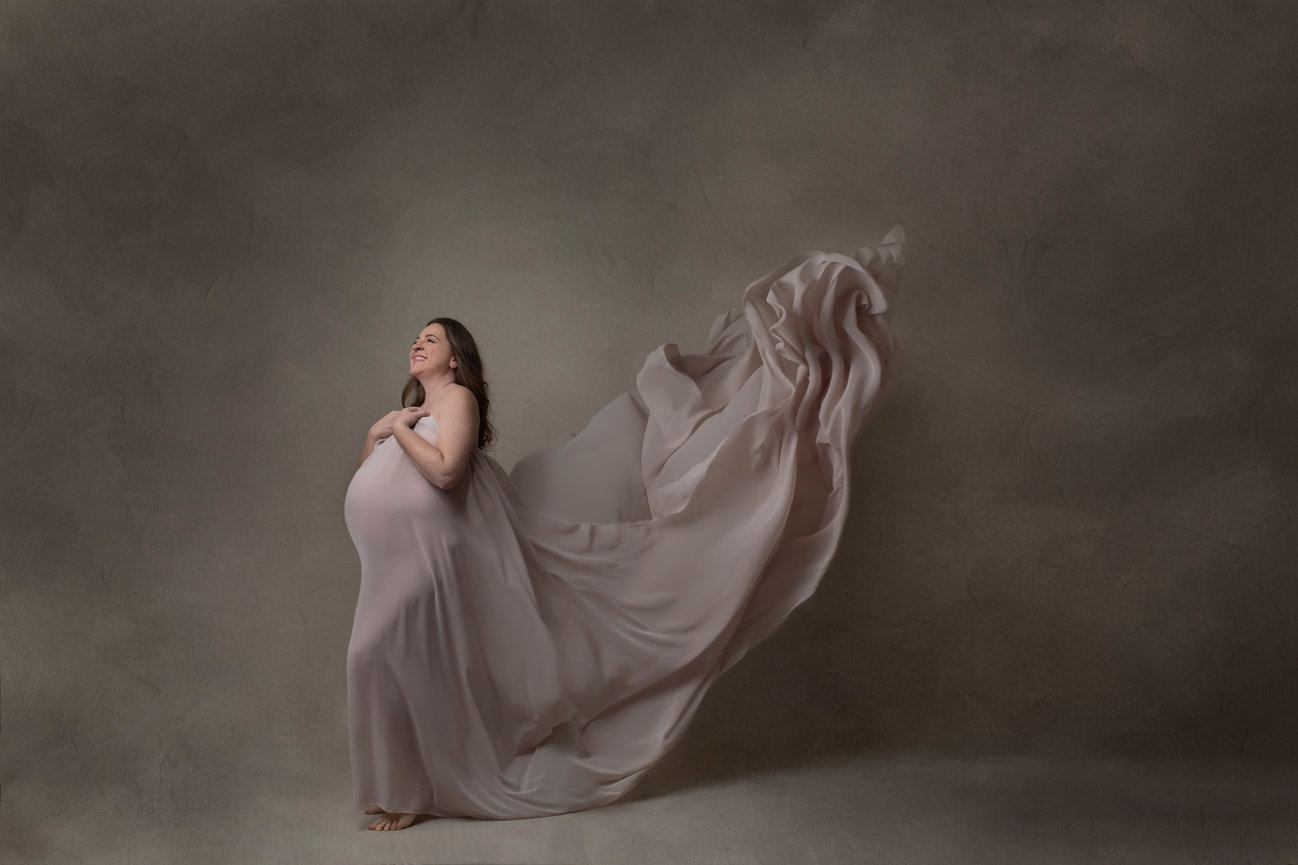 Elegant photo of older pregnant woman in her 40s forties feeling beautiful in a blowing silk fabric drape, as she gazes upward with a smile. Photo captured in the studio.