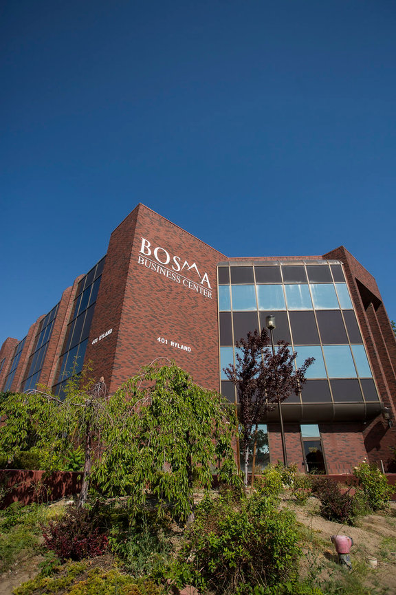 Commercial real estate and branding photo of multi story office complex exterior street view of building rising against a deep blue cloudless sky with shrubbery in the foreground.