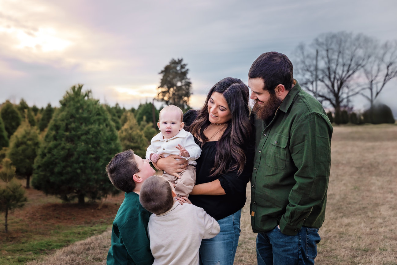 Outdoor family photography session with a family of five, with parents looking at their three children near trees under a cloudy sky.