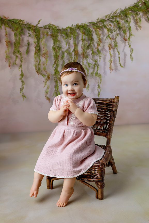 Baby girl in a pink dress sitting on a wicker chair with a floral backdrop in Kimberly A King Photography's Studio in Warsaw, VA