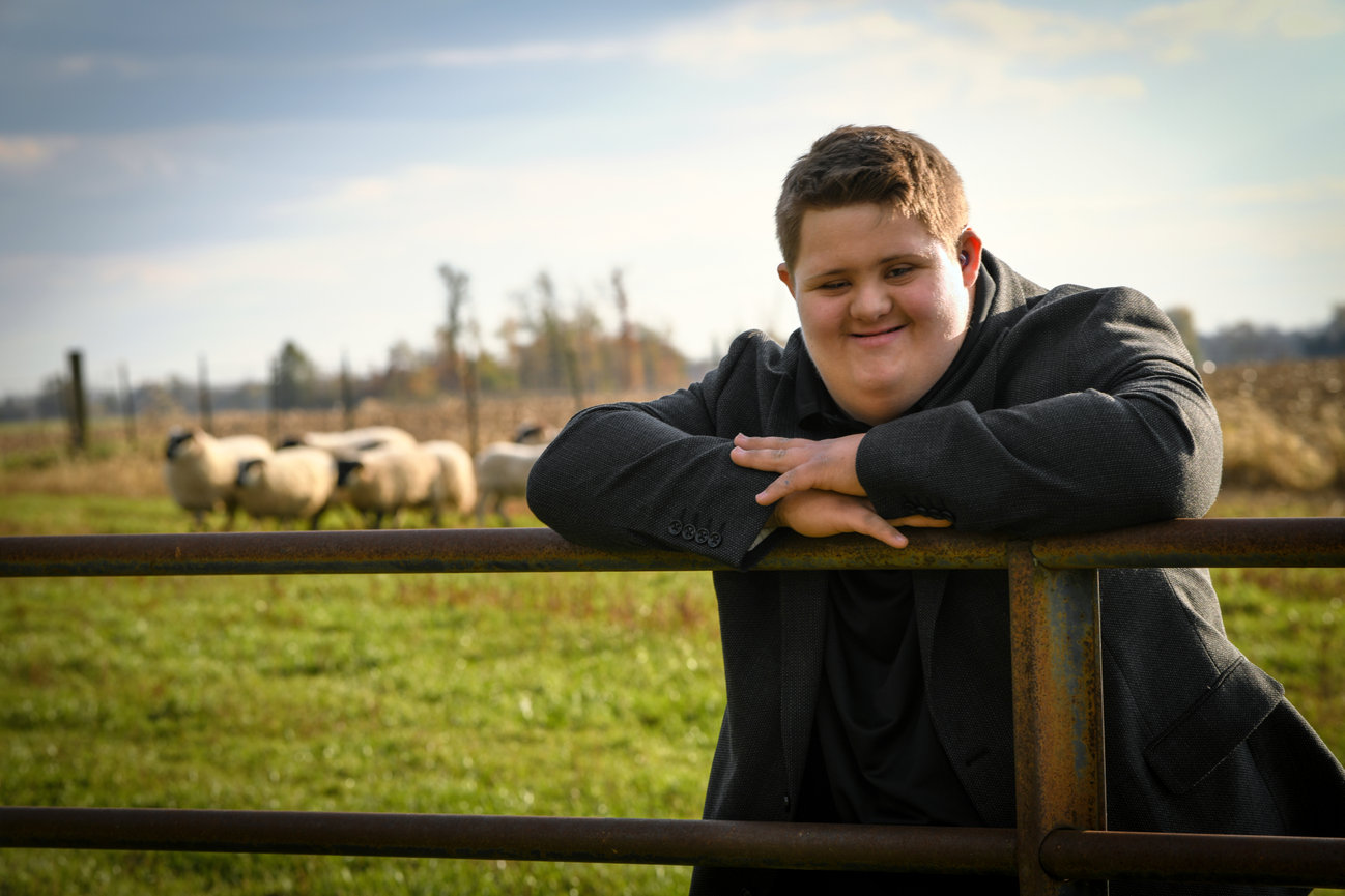 Teenage boy posing against an iron fence at a farm with sheep in the background for senior pictures in Dublin OH.