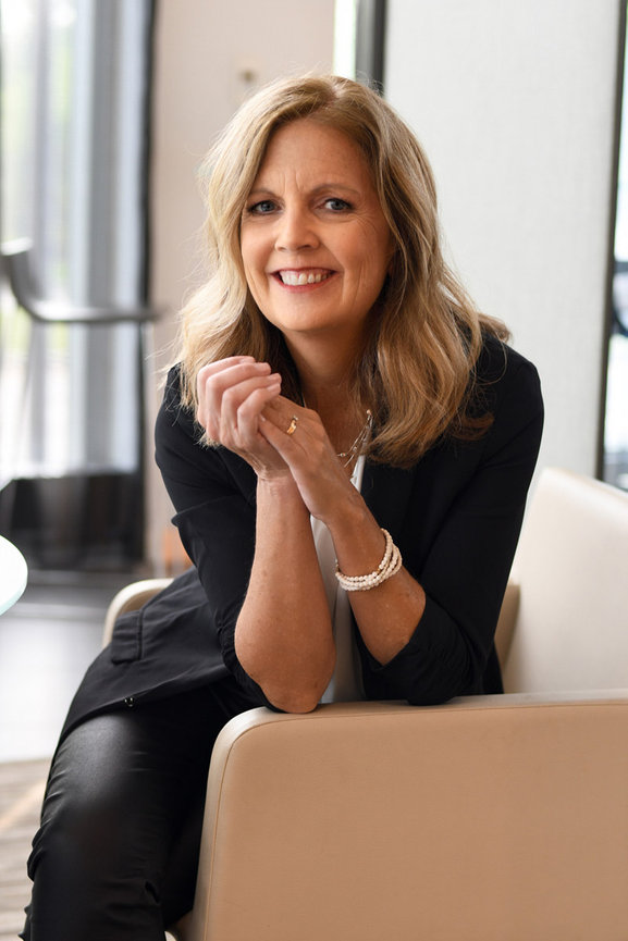 Smiling woman with medium blonde hair seated on a couch, resting her arms while wearing a black blazer and pearl accessories with headshot photographer Claudine Kosier Photography