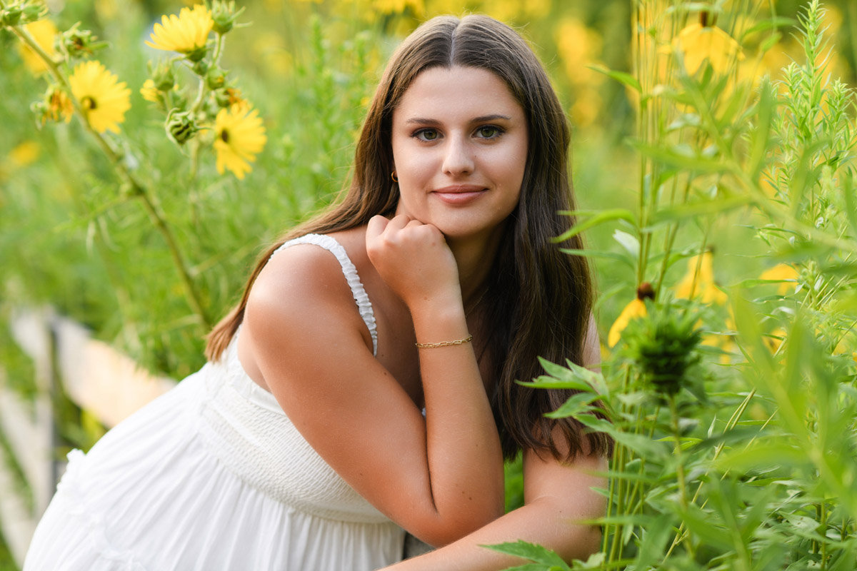 Teen girl in a white dress leaning against a wooden fence surrounded by tall yellow flowers in a vibrant garden