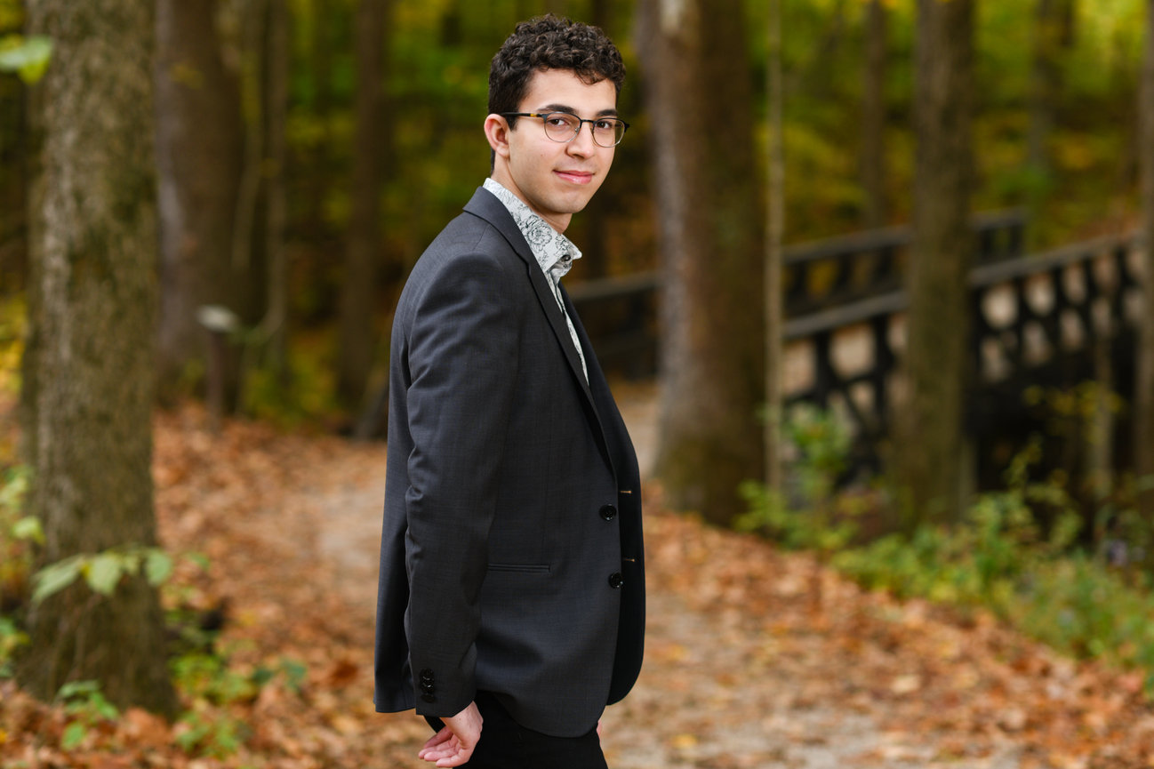 Dark haired teenage boy in a suit jacket posing against a fall backdrop for senior pictures in Dublin OH.