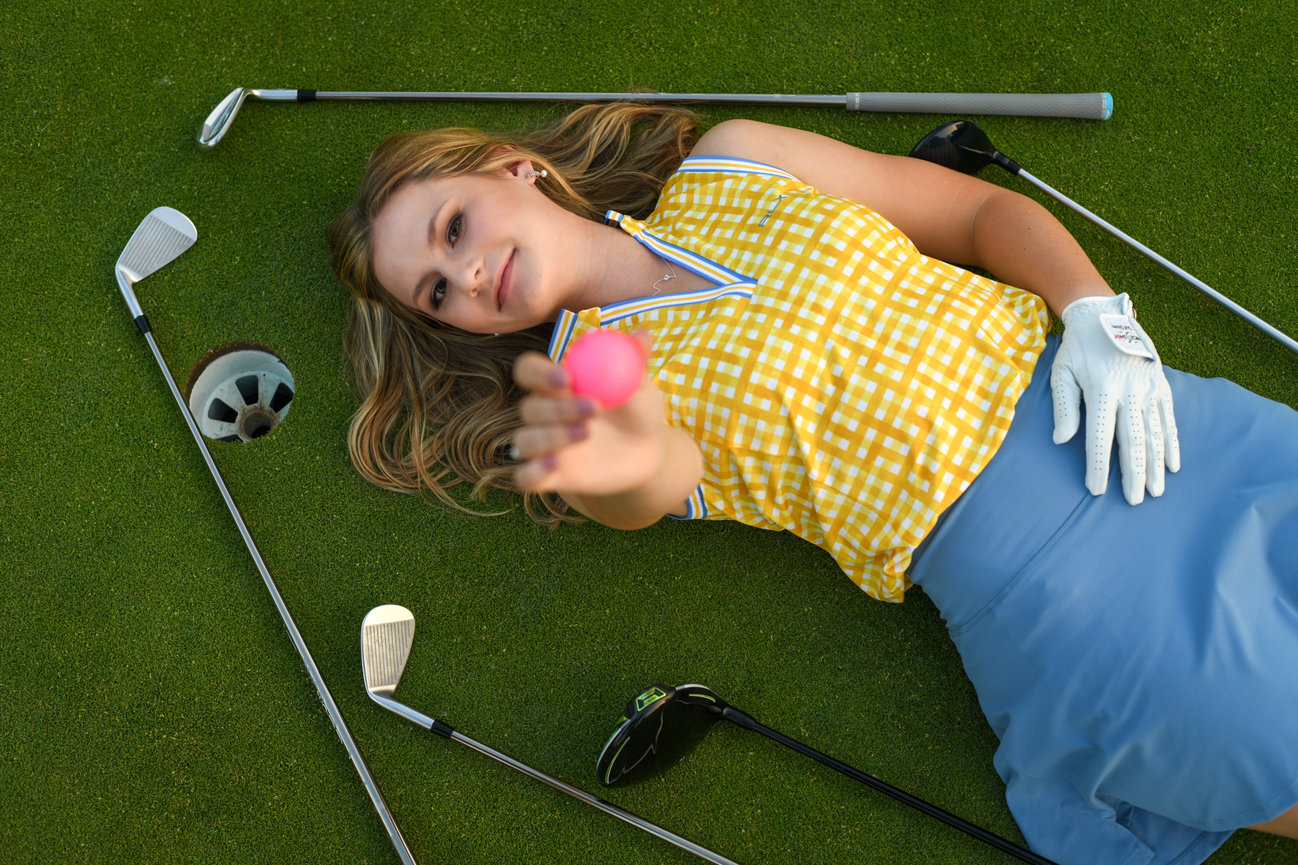Woman in a yellow checkered top lies on golf green, surrounded by golf clubs, holding a pink golf ball.