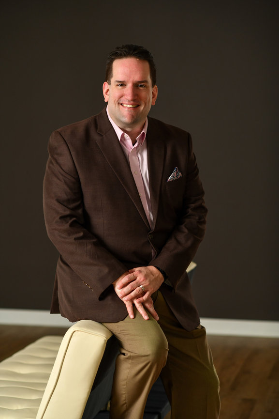 Man in a brown blazer and pink shirt poses confidently, leaning on a beige bench in a simple interior setting for his updated branding business portraits with columbus ohio branding photographer.
