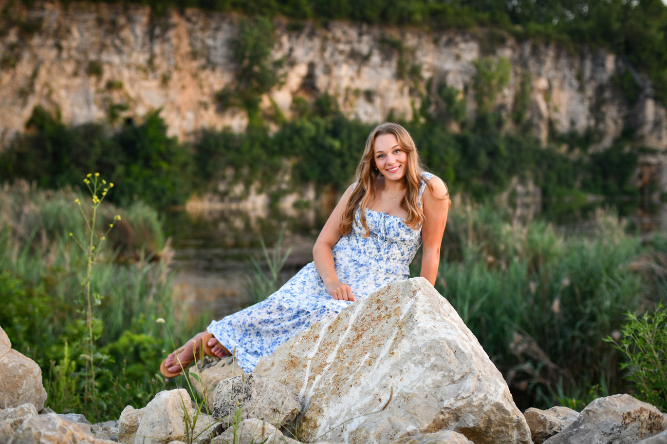 Teenage girl with long dirty blonde hair in a blue and white floral dress perched on a rock with greenery for Senior pictures in Columbus, OH.
