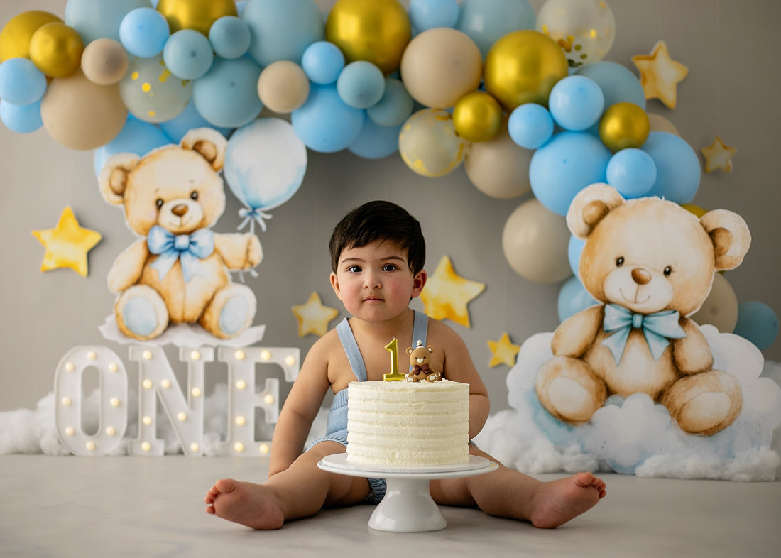 Baby boy in blue romper sitting in front of a white cake during a teddy bear–themed cake smash with blue and gold balloons by Cake Smash by Chris in Point Cook.