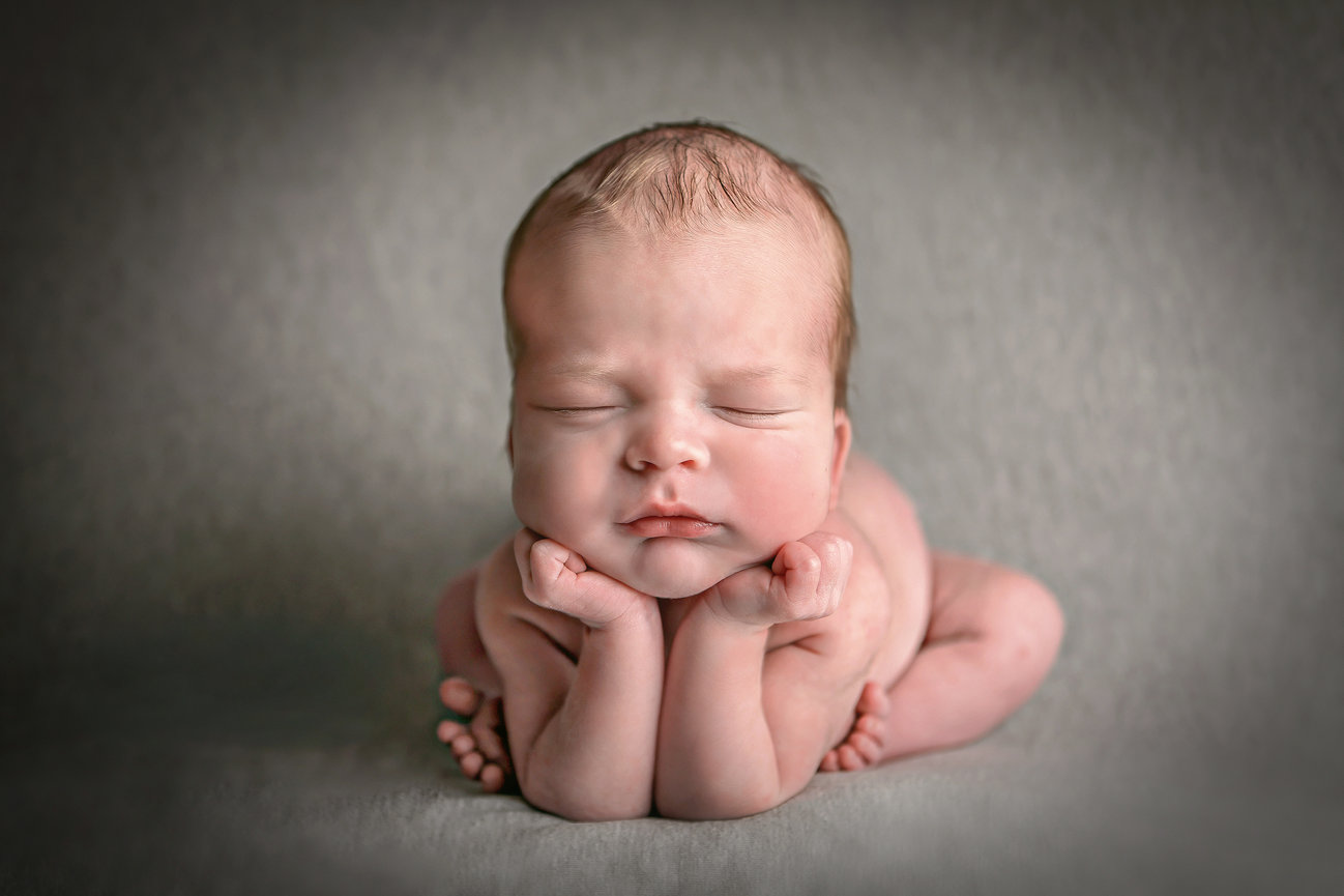 Newborn baby asleep, resting chin on hands, on a soft gray background.