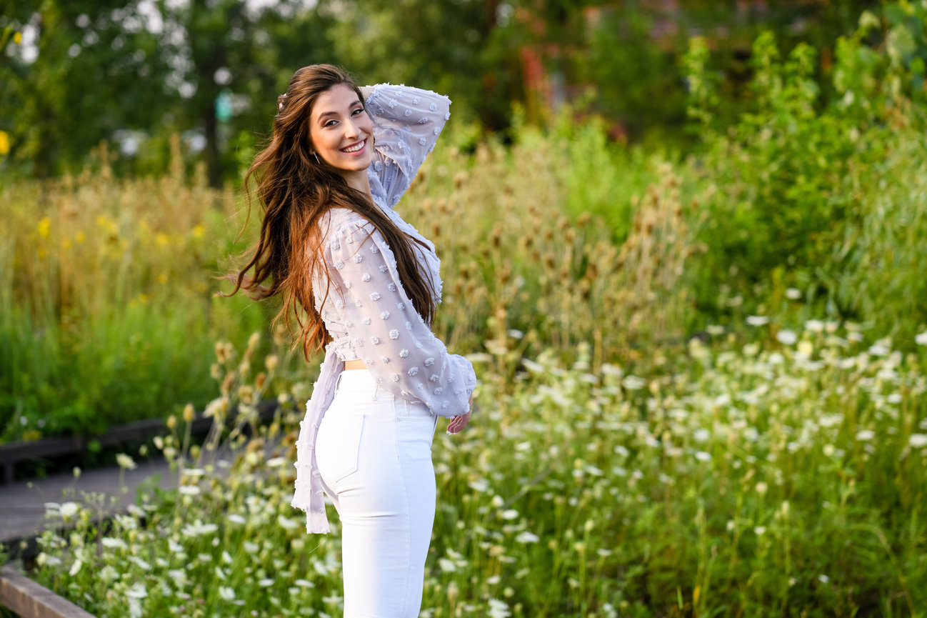 Teen girl smiling in a garden, wearing a light blouse and white pants, surrounded by greenery and wildflowers.
